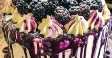 White chocolate blackberry poke cake topped with fresh blackberries, creamy frosting, and glossy blackberry ganache dripping over the sides on a cake stand.