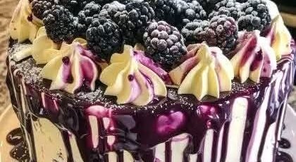 White chocolate blackberry poke cake topped with fresh blackberries, creamy frosting, and glossy blackberry ganache dripping over the sides on a cake stand.