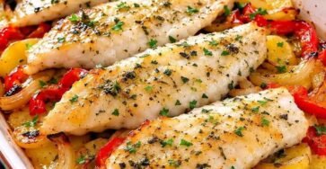 Oven-baked hake fillets served over roasted potatoes, red bell peppers, onions, garlic, and fresh parsley in a baking dish.