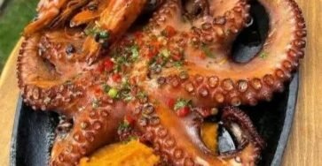 Grilled octopus with garlic shrimp served over creamy sweet potato mash alongside crispy fried plantains and grilled lemon, garnished with fresh parsley and chili.