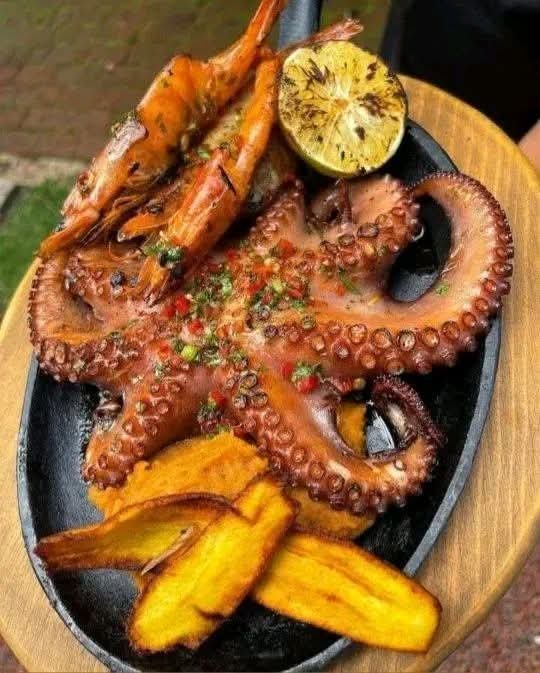 Grilled octopus with garlic shrimp served over creamy sweet potato mash alongside crispy fried plantains and grilled lemon, garnished with fresh parsley and chili.